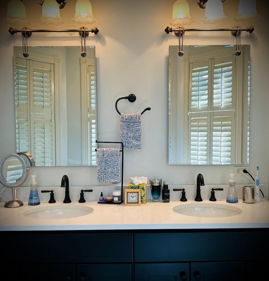 Elegant bathroom with dual sinks, dark blue vanity, white countertops, black fixtures, and white shutters above twin mirrors with sconces
