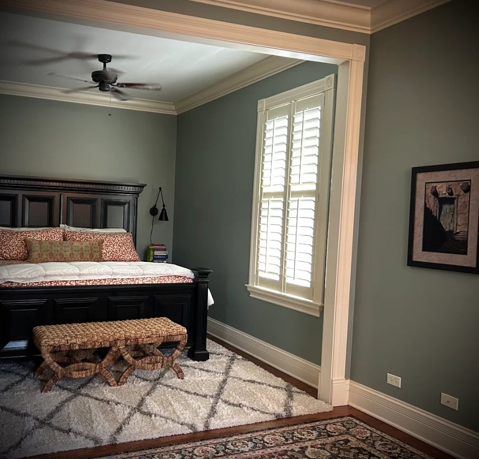 Modern bedroom with green walls, black bed with patterned pillows, woven bench at foot of bed, ceiling fan, and white shutters on tall windows