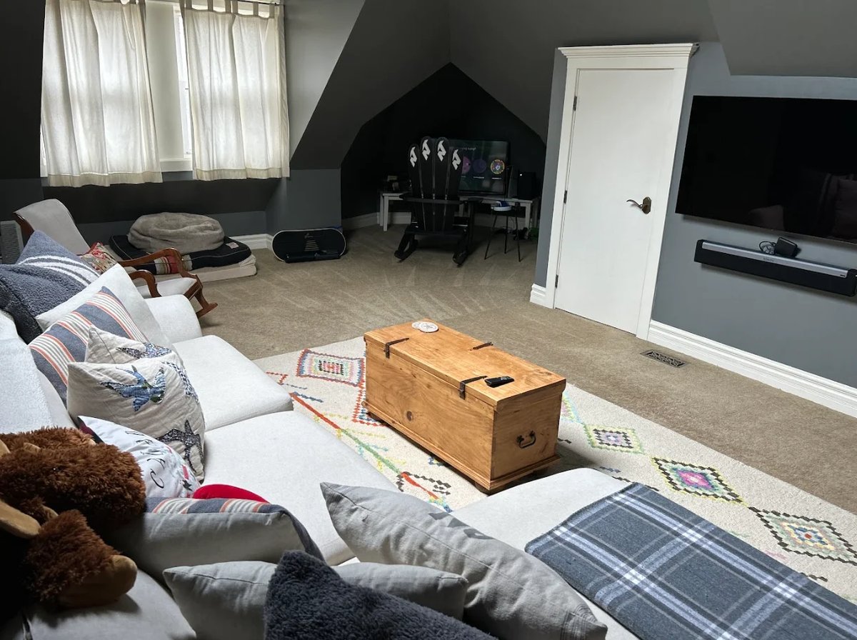 Spacious attic bedroom with sloped ceilings, white bed with pillows, wooden trunk, TV, and window with curtains