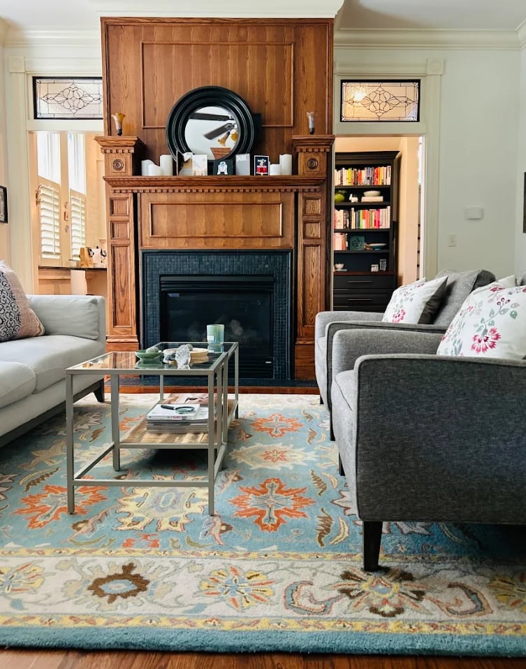 Elegant living room with wood fireplace mantel, upholstered gray furniture, patterned area rug, and bookshelf