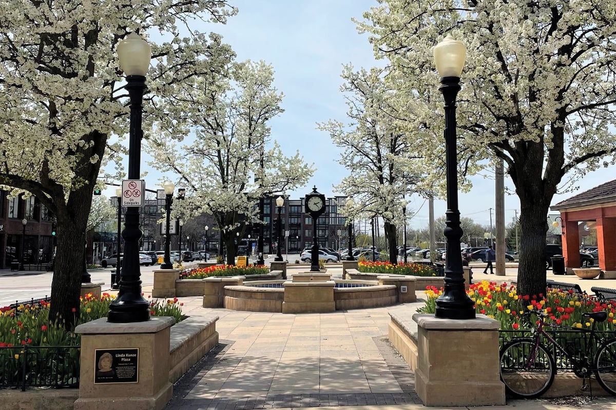 Spring town square with blooming trees, street lamps, circular stone planter, and brick buildings under clear blue sky